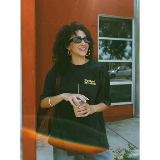 Woman wearing a black t-shirt with yellow text, holding a drink, standing in front of a red building.