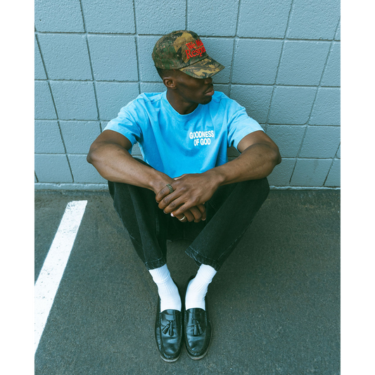 Person wearing a blue t-shirt and camouflage cap sitting against a light blue tiled wall.